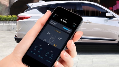 Gros plan d’une main tenant un téléphone sur l’écran duquel l’application AcuraLink est ouverte. En arrière-plan, un Acura RDX de couleur blanche est garé devant un immeuble à l’architecture moderne.