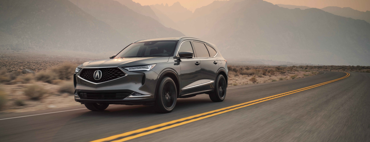 3/4 view of a grey Acura MDX driving down a road in a mountainous desert landscape. / Vue 3/4 d’un MDX gris circulant sur une route dans un paysage montagneux désert.