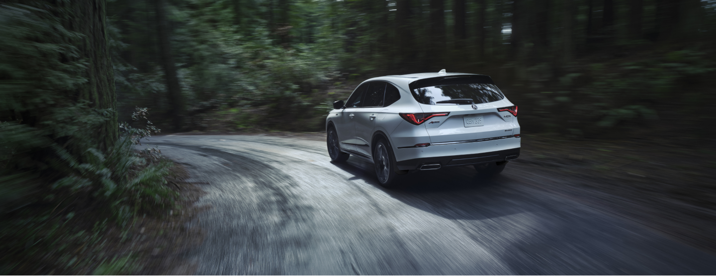Plan de trois quarts arrière d’un MDX blanc prenant un virage sur une route bétonnée étroite dans une forêt pluviale tempérée.