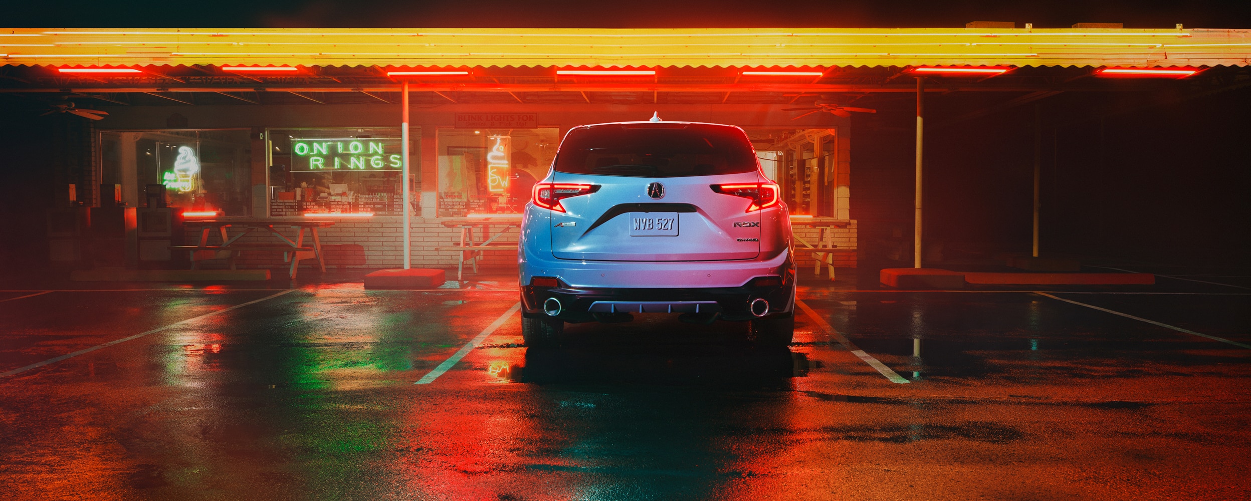 Vue arrière d’un RDX gris stationné devant un restaurant illuminé de néons, la nuit.