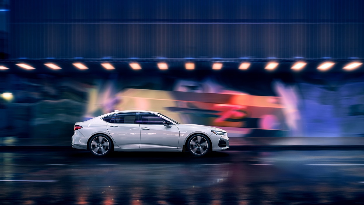 Side view of a white TLX driving down a city street at night.