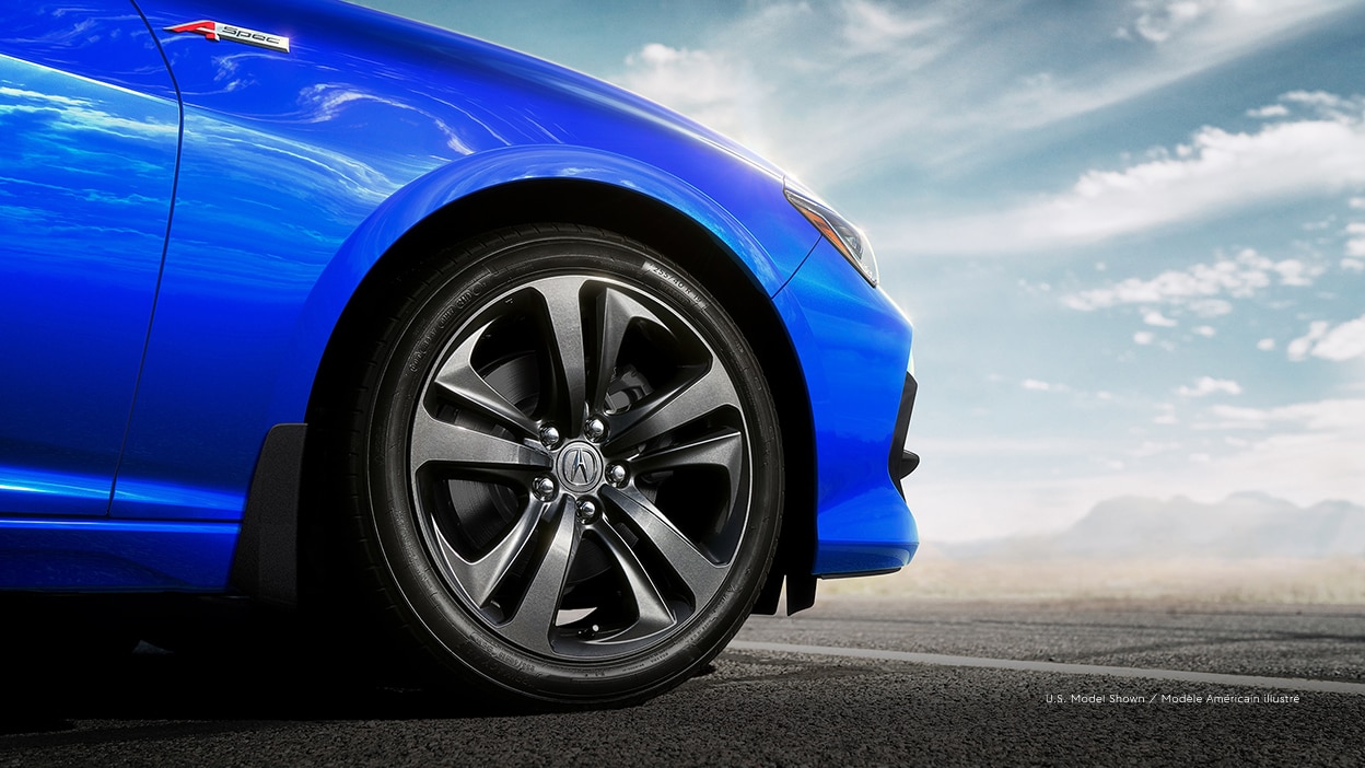 The front wheel of Acura TLX in the foreground. Mountains, clouds and blue sky in the background.