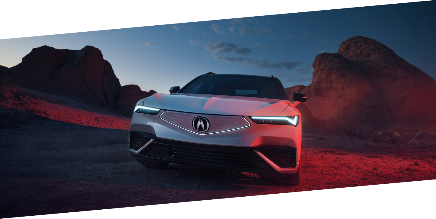 Medium wide front view of white ZDX parked in the desert at dusk. Its headlights are on and the diamond pentagon front fascia is illuminated.
