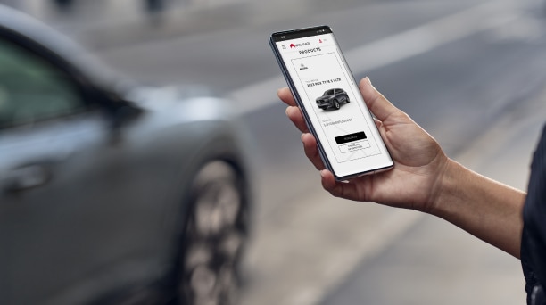A hand holds a phone displaying the MyGarage interface, with an Acura vehicle in the background.