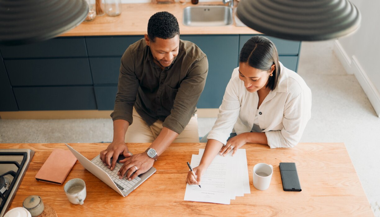 Two adults sit on a couch, smiling and looking at paperwork.