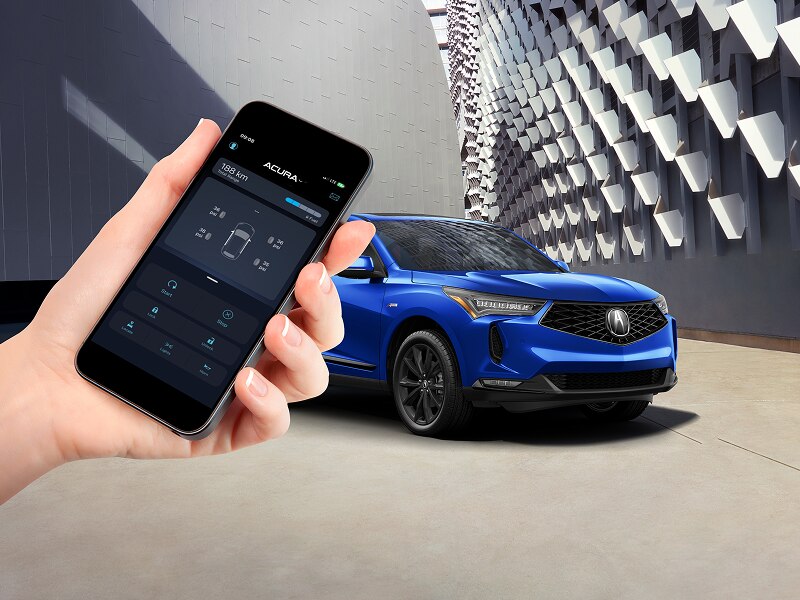 Side view of a blue Acura vehicle with a hand holding a phone in the foreground. The Acuralink™ interface appears on the screen.