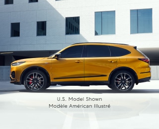 Wide side view of gold MDX Type S parked in a parking lot outside a modern, minimalistic white building.