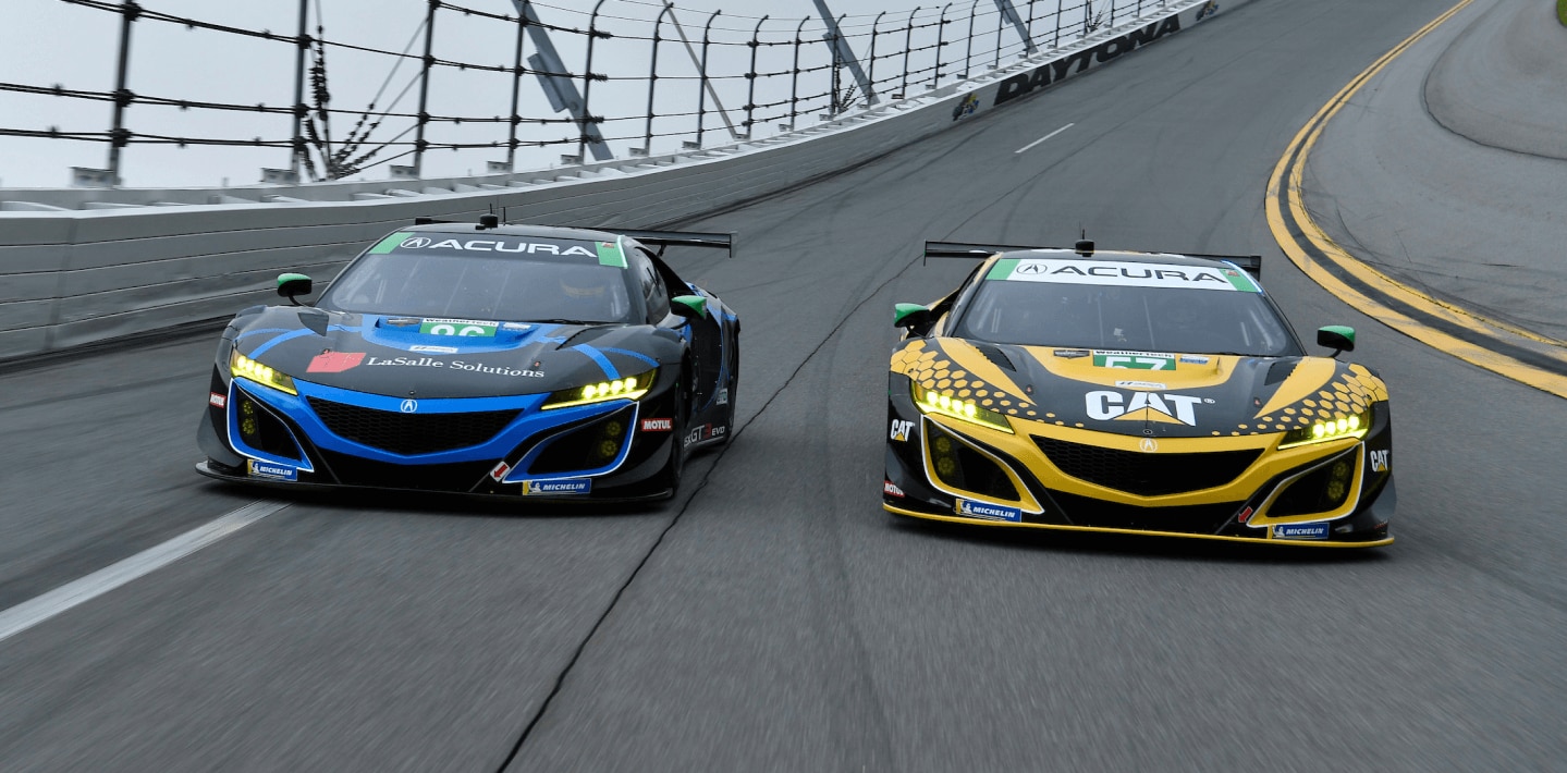 Front view of two Acura NSX GT3s, one blue and one red, both covered in sponsorship wraps, racing head-to-head on a track.  	Vue avant de deux Acura NSX GT3, l’une bleue et l’autre rouge, recouvertes de logos de commanditaires coursant sur un circuit de course.