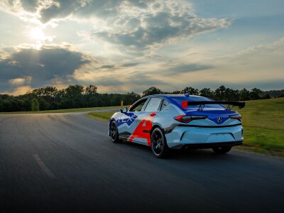 Rear, driver side angled view of an Acura TLX with red and blue racing-inspired wrap decal, driving around a track situated in an open field with trees in the distance.	Vue arrière latérale du côté conducteur d’une Acura TLX recouverte d’une décalcomanie rouge et bleue roulant sur un circuit situé dans un champ avec des arbres au loin.