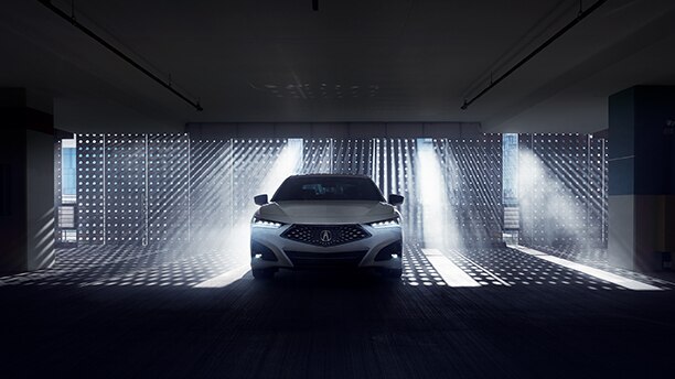 Front view of a white Acura TLX in a darkened garage with natural light shining through a gated wall behind. // Vue avant d’une Acura TLX blanche dans un garage sombre devant un mur qui laisse passer la lumière naturelle.