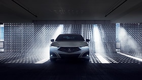 Front view of a white Acura TLX in a darkened garage with natural light shining through a gated wall behind. // Vue avant d’une Acura TLX blanche dans un garage sombre devant un mur qui laisse passer la lumière naturelle.