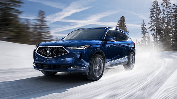 Front side-angled view of a blue Acura MDX driving a wintery road with trees and sky blurred in the background. Vue latérale avant et en angle d’un Acura MDX bleu qui roule sur une route enneigée, avec le ciel et les arbres aux contours flous en arrière-plan.