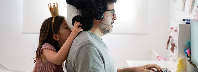 A man works at a computer while his daughter plays around behind him. / Un homme travaille devant un ordinateur pendant que sa fille joue derrière lui.