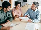 Three people sit around a table discussing paperwork. / Trois personnes sont assises autour d'une table pour discuter au sujet de documents.