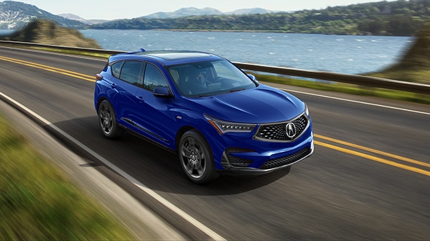 Vue latérale de face d’une Acura bleue roulant sur une route pavée le long d’une étendue d’eau et d’une colline.