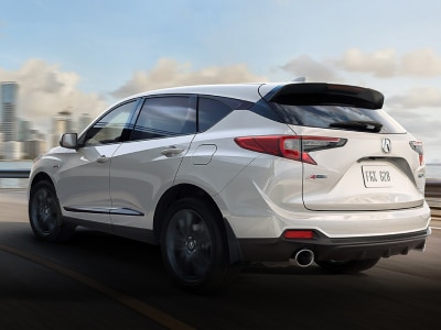 Rear left image of an Acura RDX that emphasises the spoiler and aesthetics of the vehicle while driving fast on the road causing the buildings in the background to blur. // Image arrière gauche d'un Acura RDX qui met l'accent sur le becquet et l'esthétique du véhicule tout en conduisant rapidement sur la route, ce qui rend les bâtiments à l'arrière-plan flous