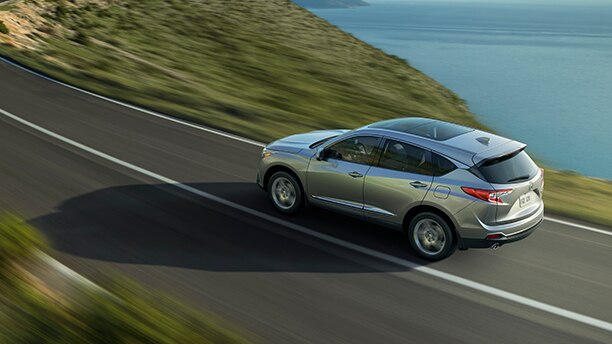 Rear-facing side view of silver Acura driving down a paved road alongside a body of water and hillside. // Vue latérale arrière d’une Acura grise métallisée roulant sur une route pavée le long d’une étendue d’eau et d’une colline.