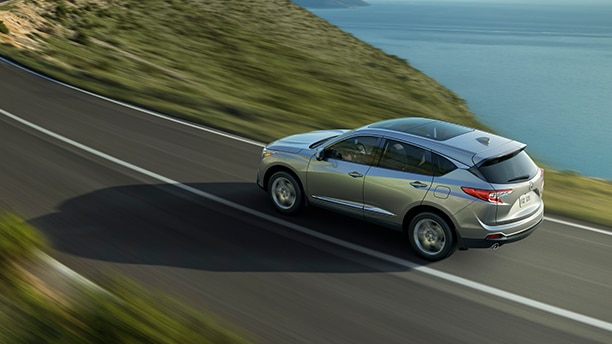 Rear-facing side view of silver Acura driving down a paved road alongside a body of water and hillside. 	Vue latérale arrière d’une Acura gris métalisée roulant sur une route pavée le long d’une étendue d’eau et d’une colline.