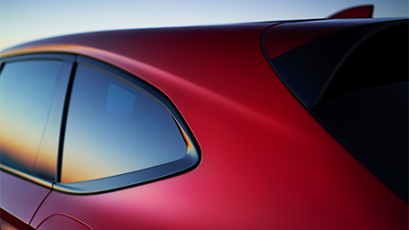 Zoomed-in side-view of the back of a red Acura, with sunset reflections on the windows. // Vue latérale agrandie de l'arrière d’une Acura rouge, avec des reflets de coucher de soleil sur les vitres.