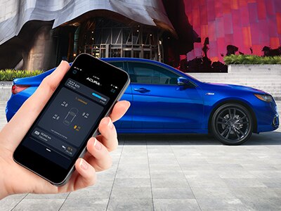 Shot of a hand holding a smart phone displaying the AcuraLink™ app in the foreground of an image. The background image features a blue Acura ILX parked in front of a futuristic building./Prise de vue d'une main tenant un téléphone intelligent affichant l'application AcuraLinkMC au premier plan d'une image. L'image de fond présente une Acura ILX bleue stationnée devant un bâtiment futuriste.
