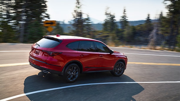 Passenger side view of a red 2022 Acura MDX driving around a bend on tree-lined paved road. // Vue latérale du côté passager d'un Acura MDX 2022 rouge roulant dans un virage sur une route pavée bordée d'arbres.